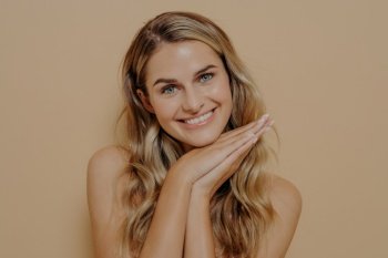 Smiling merry blonde young woman with long wavy hair feeling happy and showing it with gesture, resting head on hands together under chin, isolated in front of orange background. Calm blonde teenage girl smiling and looking straight at camera