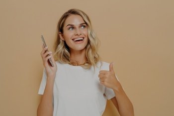 Young satisfied girl in casual clothes holding mobile phone and showing thumb up gesture, being excited to hear positive news while standing isolated over brown background, looking upside with smile. Young satisfied girl in casual clothes holding mobile phone and showing thumb up gesture, being excited