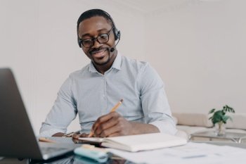 African american man wearing headset working distantly on computer at home office. Black guy student in headphones learns online at laptop, takes notes. E-learning, distance education.. African american man in headset learns online at laptop, takes notes. Elearning, distance education
