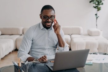 Smiling african american businessman talking on phone looking at laptop at office desk. Black male businessperson marketing and sales manager advising client, makes offer, sells, conducts negotiation. African american businessman talking on phone at laptop advises client, sells, conducts negotiations