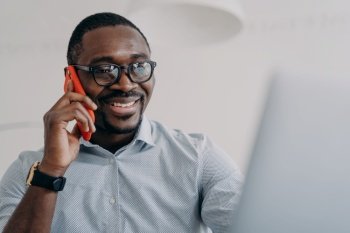 Successful african american businessperson in glasses hold mobile phone makes nice business call sitting in front of laptop. Black male friendly manager consulting a client, talking on smartphone.. Happy african american businessperson in glasses hold phone makes business call in front of laptop