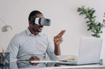 African american businessman wearing vr glasses works in augmented reality at laptop. Black young male office worker immersed in virtual reality, using innovation technology, managing business project. African businessman wearing vr glasses works at laptop manages business project in virtual reality
