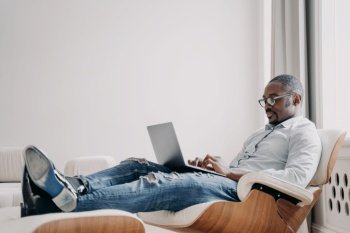 Focused african american man freelancer working online at laptop, typing email or message, sitting in modern comfortable armchair. Black male businessperson developing business project. Remote work. African american man working online on business project at laptop, sitting in armchair. Remote job