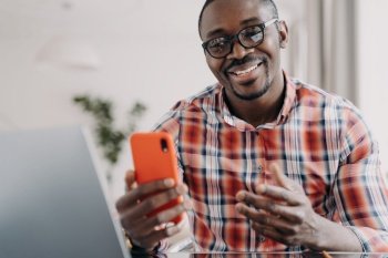 Happy african american man wearing glasses holding phone using modern apps sitting at laptop. Smiling young black guy looking at smartphone, reading good news message, chatting online.. Happy african american man wearing glasses holding smartphone uses modern apps sitting at laptop