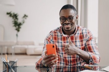 Happy african american man showing like thumb up hand, holding smartphone smiling. Pleased black guy looking at phone screen, showing positive feedback gesture, chatting by video call, sitting at desk. African american man shows thumb up hand gesture, holding phone smiling. Positive feedback gesture