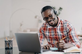 African american student young man getting remote education at laptop. Happy black male wearing headset listening lecture, taking notes while online lesson by video conference. Distance learning . African american student in headset learning at laptop online, makes notes. Distance education