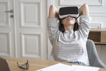 Young happy female employee in VR goggles at workplace watching movies, relaxed woman office worker sitting at desk wearing virtual reality helmet and enjoying 3D experience at work. Young happy female employee in VR goggles at workplace watching movies