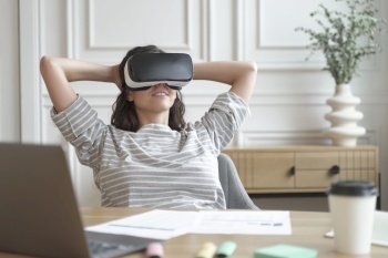Relaxed young woman office worker wearing VR headset or helmet watching in 360 degrees video or movie during break at work, sitting in 3d glasses at workplace with hands behind head.. Relaxed young woman office worker in VR headset or helmet watching in 360 degrees video or movie