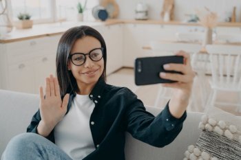 Smiling young female greeting friend, waving hand, having video conversation by smartphone sitting on couch at home. Businesswoman wearing glasses chatting by videocall make hello gesture, welcoming.. Smiling female holding phone, greeting, waving hand, chatting by video call sitting on couch at home