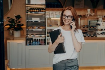 Woman entrepreneur with ginger hair has telephone conversation holds laptop computer discusses time of meeting with colleague poses indoor at own shop wears spectacles for vision correction.. Woman entrepreneur with ginger hair has telephone conversation holds laptop