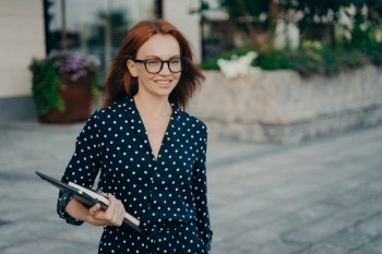 Successful redhead female office worker returns from work poses outside with organizer modern gadgets wears spectacles black polka dot dress focused into distance strolls at street. Business people. Female office worker poses outside with organizer modern gadgets