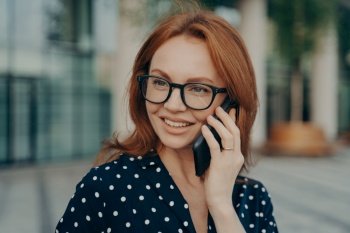 Photo of beautiful redhead woman has telephone talk uses smartphone for call looks away wears spectacles polka dot clothes poses against blurred background outdoor phones via mobile application. Beautiful redhead woman has telephone talk uses smartphone for call