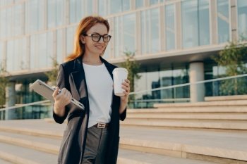 Beautiful stylish redhead businesswoman with laptop drinking coffee from disposable paper cup while going to work in morning, female business professional walking outdoors on city street and smiling. Stylish businesswoman with laptop drinking coffee from disposable paper cup while going at work