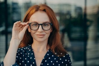 Portrait of confident beautiful red-haired business woman adjusting spectacles eyeglasses while posing on city street in summer, looking at camera with confidence, blurred urban background. Confident beautiful red-haired business woman adjusting spectacles eyeglasses posing on street