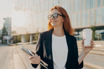 Overjoyed happy ginger woman in spectacles drinking coffee and holding laptop while walking outdoors on sunny day, business woman enjoying her way to office in morning, smiling and looking asidee. Overjoyed happy ginger woman in spectacles drinking coffee and holding laptop while walking outdoors