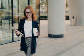 Positive redhead young European woman in elegant clothes holds notebook pen and laptop drinks aromatic coffee from paper cup walks outdoors near office building being in hurry for formal meeting. Positive redhead young European woman in elegant clothes holds notebook pen and laptop
