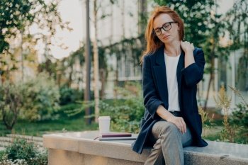 Frustrated redhead young woman feels very tired suffers from neck pain sits in park outdoors wears transparent glasses and formal wear focused down thinks about something fatigue after work.. Frustrated redhead young woman feels very tired suffers from neck pain sits in park