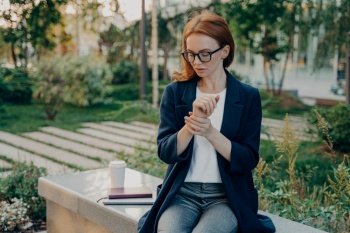 Young tired redhead businesswoman measuring pulse on wrist, checking heart rate after stressful working day in office, overworked female sitting in park and relaxing, feeling exhausted. Healthcare. Young tired redhead business woman measuring pulse on wrist, checking heart rate, sitting in park