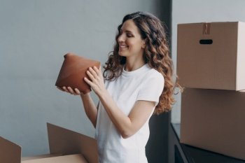 Relocation and delivery service concept. Happy girl unpacking cardboard boxes and holding the vase. Woman in casual outfit. Smiling attractive spanish woman packing boxes to move.. Relocation and delivery service concept. Happy girl unpacking cardboard boxes and holding the vase.