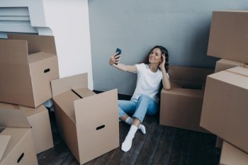 Home owner is taking selfie in new luxurious apartment. Happy woman unpacking cardboard boxes. Girl is taking photo on phone and sitting on the floor. Independence and success concept.. Happy woman unpacking boxes and taking selfie in new luxurious apartment. Success concept.