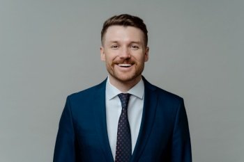 Portrait of cheerful successful happy businessman or male ceo executive in formal wear suit smiling at camera and looking pleased after successful deal while posing against grey studio background. Happy professional businessman in suit smiling at camera