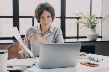 Focused Italian businesswoman making video call in office, holding document with financial data looking at laptop screen, proffesional female real estate agent leading remote meeting with client. Serious focused Italian businesswoman making video call on laptop in office