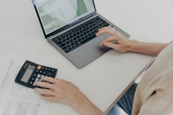 Cropped shot of unrecognizable woman sits at desk with laptop computer calculator and papers calculates montly expenses studies utility bills counts income uses e banking application plans investments. Unrecognizable woman sits at desk with laptop computer calculator papers calculates montly expenses