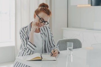 Happy beautiful redhead European woman makes notes in personal planner smiles gently holds pen poses at desktop drinks fresh pure water poses in spacious living room writes memo in her diary.. European woman makes notes in personal planner smiles gently holds pen poses at desktop