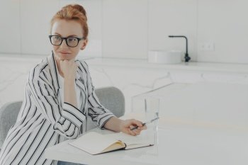 Thoughtful redhead woman distracted from work makes notes in notepad holds pen wears spectacles striped shirt poses at white desktop writes down information in notebook works distantly from home. Thoughtful redhead woman distracted from work makes notes in notepad holds pen