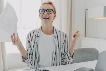 Excited young woman with paper bills in hands feeling euphoric, happy about finally paying down her mortgage debt, exclaiming yes with excitement while sitting at table with laptop. Excited young woman with paper bills in hands finally paying down her mortgage debt at home