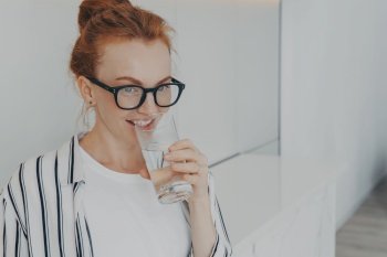 Happy healthy beautiful young redhead woman with clean skin drinking water in morning and smiling, beautiful female holding glass in hand, preventing dehydration while standing in kitchen at home. Happy healthy beautiful young redhead woman drinking water in morning at home