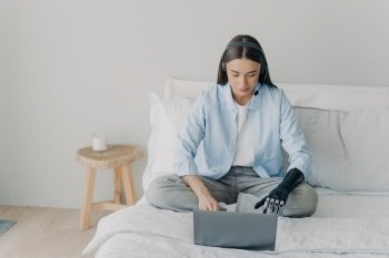 Disabled woman with artificial arm works from bedroom. Freelancer has online meeting on bed using headset. Handicapped girl assists remotely.