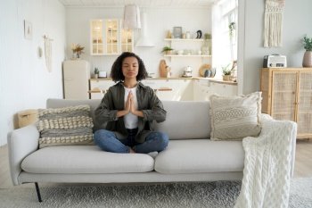 Mixed race girl embraces a healthy lifestyle, practicing yoga and meditation at home, sitting in a lotus pose on the couch.