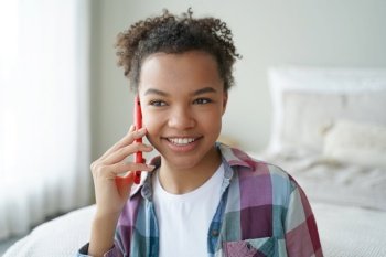 Smiling mixed race teen girl happily answers a phone call, enjoying a pleasant conversation with a friend in the comfort of her bedroom at home.