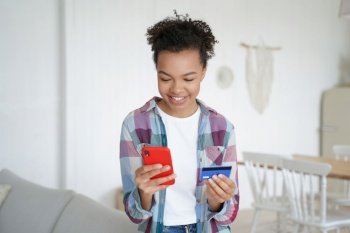 Mixed race young girl securely makes online purchases at home, confidently holding her banking credit card and utilizing e-banking phone apps.