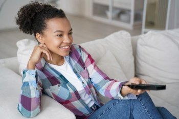 Smiling mixed race teen girl relaxes on the sofa at home, indulging in watching TV series and effortlessly changing TV channels.