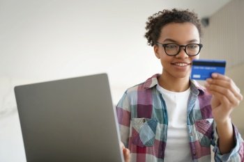 Mixed race teen girl manages her finances, confidently holding a credit card and utilizing online banking services on her laptop for e-commerce purposes at home.