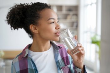 Healthy mixed race girl enjoys morning routine, drinking pure water from glass at home, embracing a healthy lifestyle.