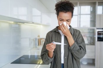 At home, a sick mixed race girl blows her nose and checks her temperature with an electronic thermometer due to flu and fever symptoms.