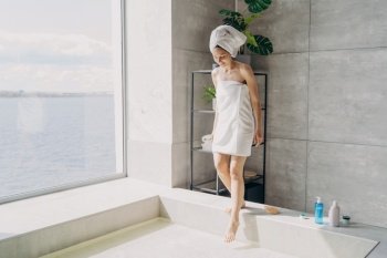 Girl in towels steps into water-filled bath in sunny modern bathroom. Self-care, spa, hygiene. 