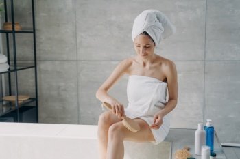 Towel-clad woman massages hip with brush in bathroom. Cellulite treatment, body care