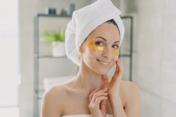 Smiling woman in towel applies golden hydrogel patches under eyes in bathroom. Morning antiaging skincare routine.