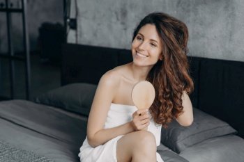 Smiling latina woman tidies long brown hair with hairbrush, sitting on bed. Morning beauty routine, haircare. Hair growth treatment ad