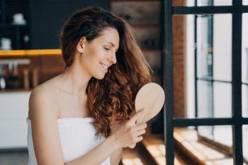 Young hispanic woman brushes long brown curly hair with wooden hairbrush after shower. Morning beauty routine at home. Haircare cosmetics ad.