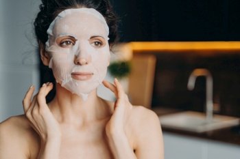 Young woman applies moisturizing rejuvenating facial sheet mask for complexion improvement at home. Skincare, daily beauty routine, self-care