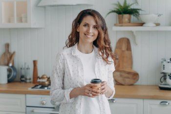 Relaxed smiling hispanic woman in cozy kitchen holds coffee, looks at camera. Pleased homeowner enjoys domestic routine at home