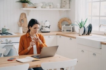 Busy woman works at home, sitting at kitchen table with laptop. Pensive housewife shops online, plans purchases, makes to-do list, learns language