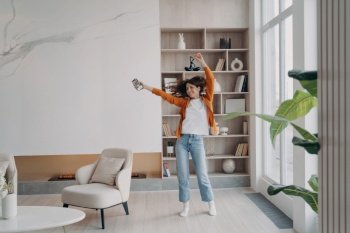 Energetic woman dances alone in living room to smartphone music. Joyful homeowner enjoys music, has fun in modern apartment.