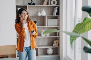 Smiling woman uses wireless earphones and mobile app at home. Positive businesswoman chats, listens to music or audio book in modern apartment.