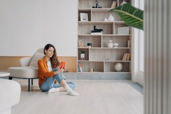 Smiling woman with wireless earbud uses phone, sitting on heated floor in living room. Online video call or music/podcast listening at home.